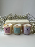 Three Scentsei Fragrance candles with labels in front of a cream luxe linen box and white flowers. A candle trio set "Her Rooted Radiance" a Mother's Day collection by Scentsei Fragrance. 